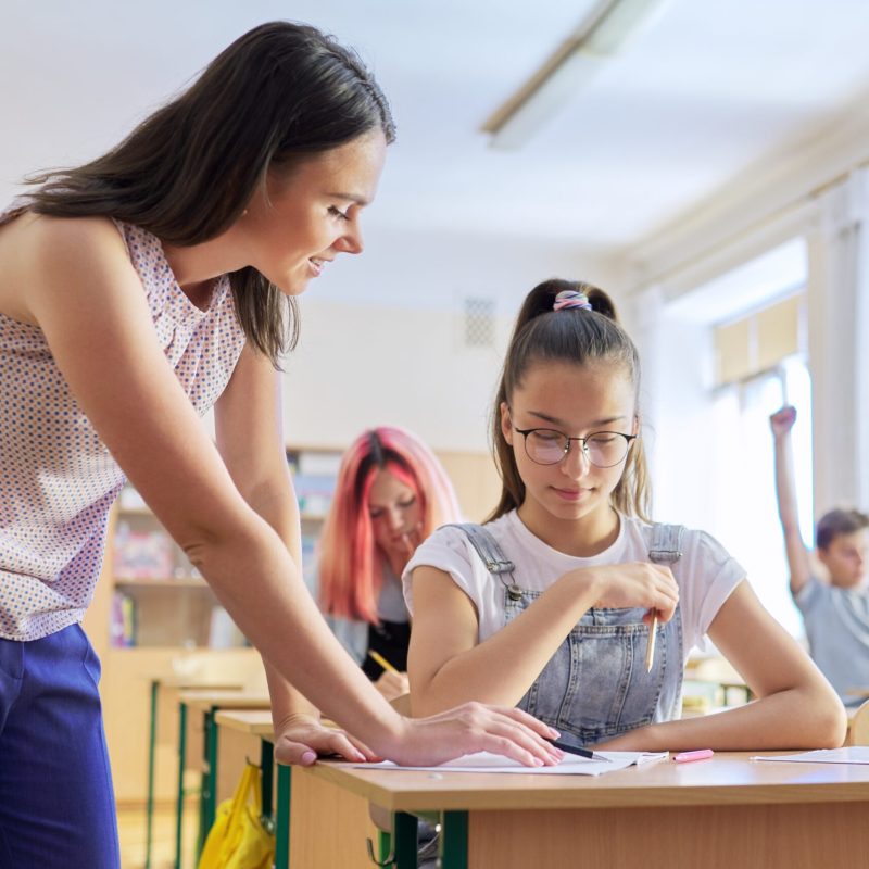 Young woman teacher teaches lesson in class of teenage children, teacher stands near school desk with girl student, checks knowledge. Education, school, college, teaching concept